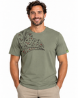 A man wearing an olive green crewneck t-shirt with a large Polynesian tribal graphic on the chest, paired with khaki pants, against a plain white background.