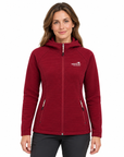 A woman wearing a textured red hooded zip-up jacket with a white embroidered logo on the left chest, paired with dark gray pants, against a plain white background.