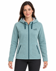 A woman wearing a light blue heather zip-up hoodie with dark teal drawstrings, zippers, and a small 'WILD AFRICA' logo on the left chest, paired with black pants, against a plain white background.