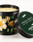 A black metal travel candle tin with a lid, shown open with cream-colored wax inside. The tin features a label with a white flower, green leaves, and a bee, while the lid is dark green with white text reading 'MANUKA HONEY travel candle' and 'wild kiwi NEW ZEALAND'.