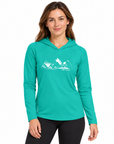 A woman modeling a teal long-sleeve hooded athletic shirt with a white mountain graphic and the words 'THE PEAK' on the chest, paired with black leggings, against a plain white background.