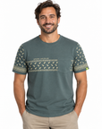 A man wearing a muted green short-sleeve t-shirt with a chest pocket and a horizontal patterned band across the chest and sleeves, paired with khaki pants, against a plain white background.