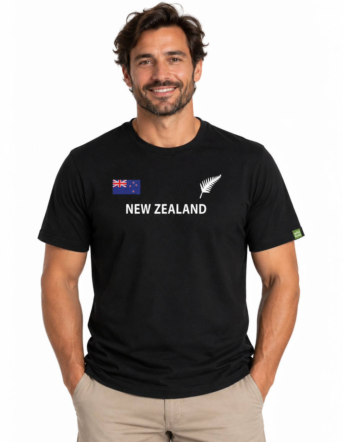 A man wearing a black short-sleeve t-shirt with 'NEW ZEALAND' printed in white across the chest, featuring the New Zealand flag and silver fern logo on the left chest, paired with khaki pants against a white studio background.
