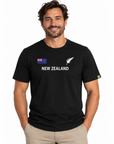 A man wearing a black short-sleeve t-shirt with 'NEW ZEALAND' printed in white across the chest, featuring the New Zealand flag and silver fern logo on the left chest, paired with khaki pants against a white studio background.