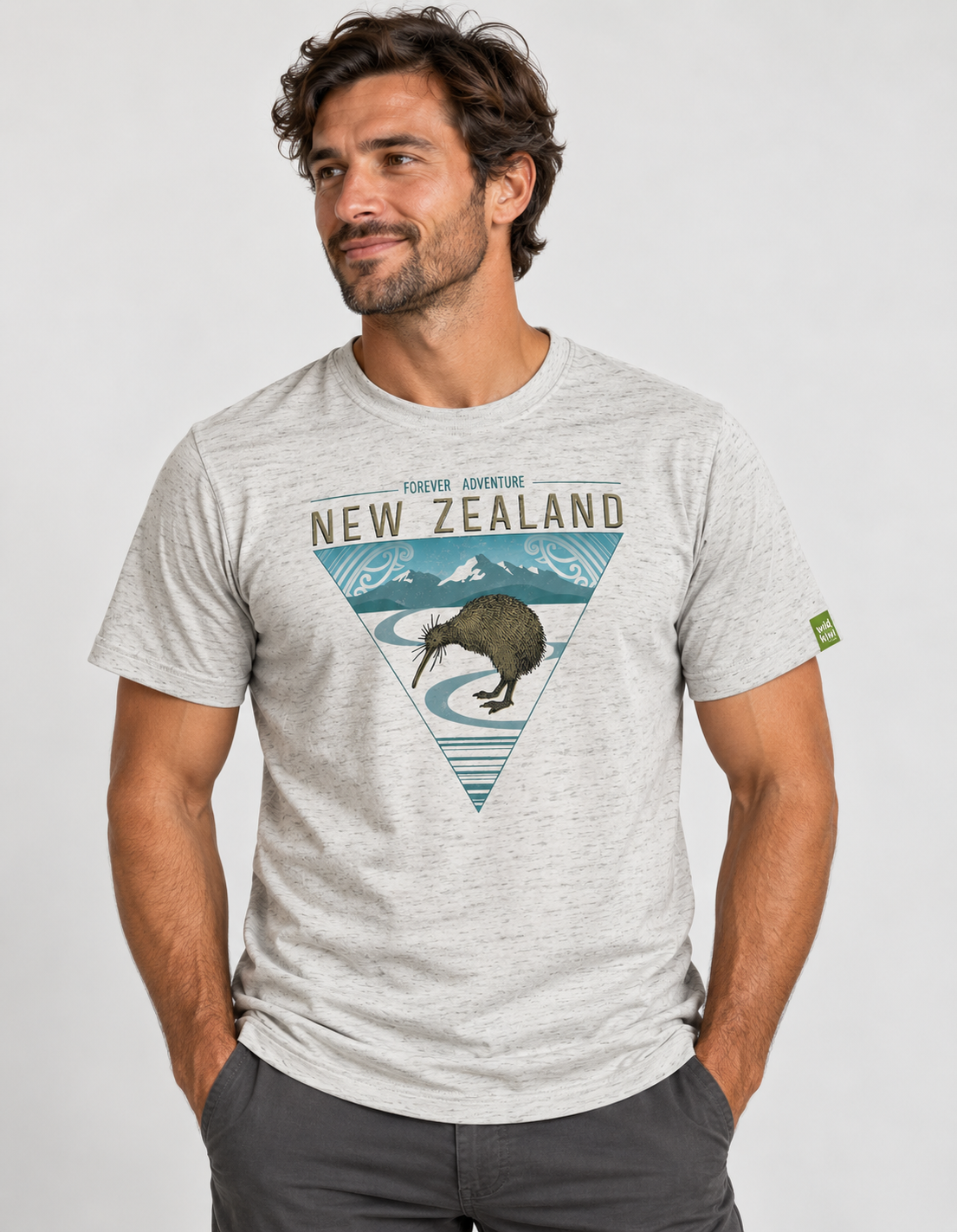 A man wearing a light heather gray t-shirt with a 'New Zealand' kiwi graphic, paired with dark gray pants, against a light gray studio background.