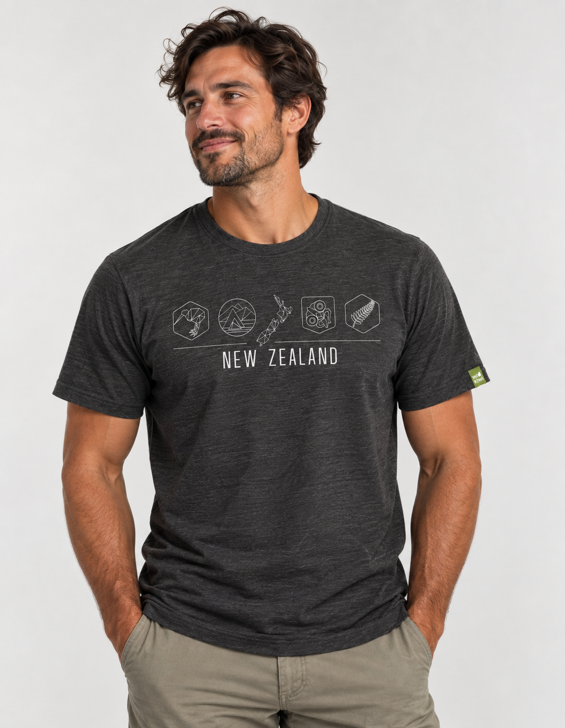 A man wearing a dark heather gray t-shirt with a white graphic of New Zealand landmarks and the text 'NEW ZEALAND' on the chest, paired with khaki pants, against a light gray background.