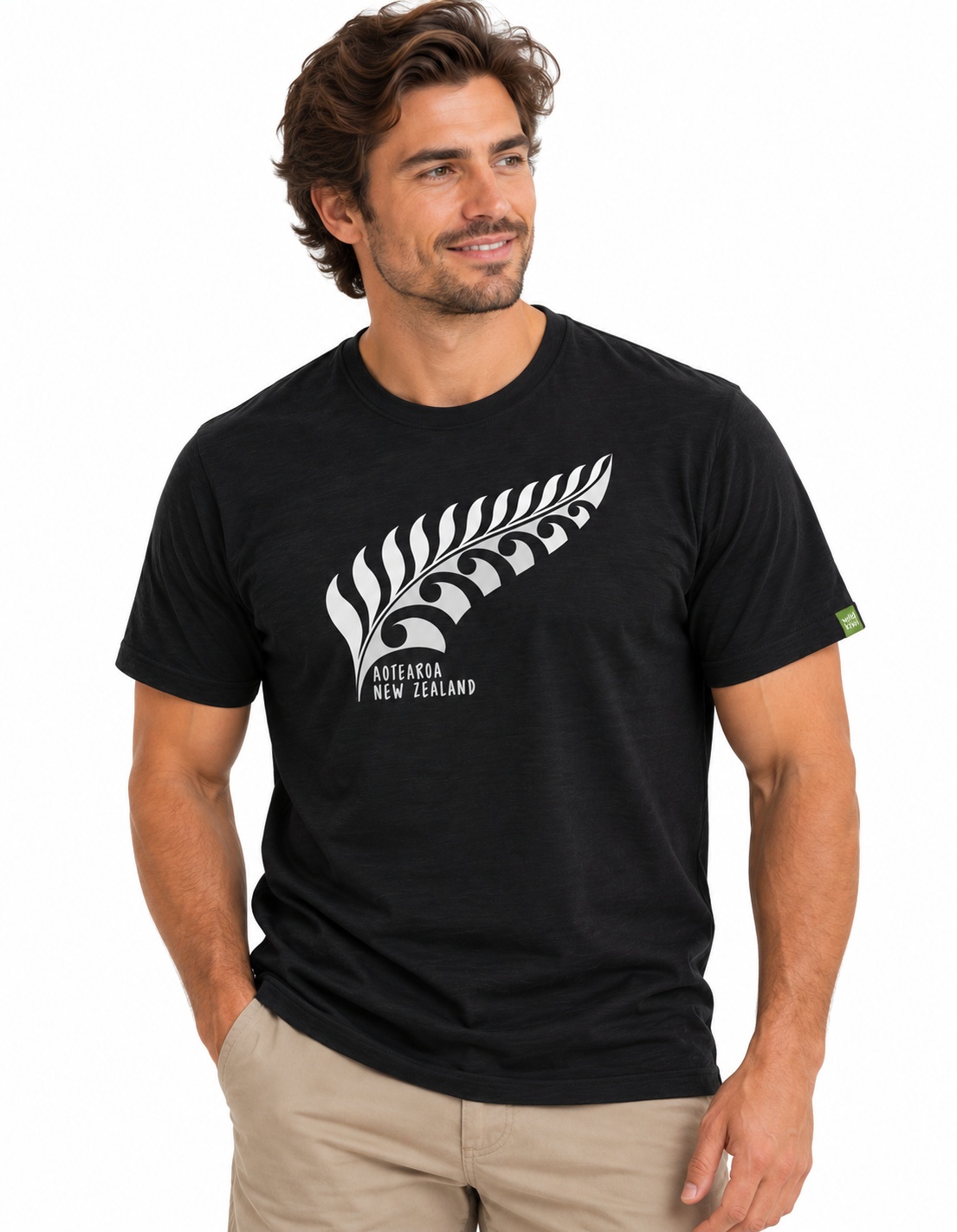 A man wearing a black short-sleeve t-shirt with a white fern leaf graphic and 'Aotearoa New Zealand' text on the chest, paired with khaki pants, against a plain white background.