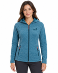 A woman modeling a heathered blue zip-up hoodie with a small embroidered logo on the left chest, set against a plain white background.