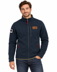 A man wearing a dark navy blue fleece jacket with a full-zip front, a brown brand patch on the chest, and red zipper pulls, paired with blue jeans against a plain white background.