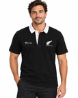 A man wearing a black short-sleeve polo shirt with a white collar. The shirt features the New Zealand fern logo on the left chest and a New Zealand XV match logo on the right chest.