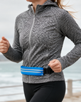 A woman running outdoors near the ocean wears a blue and white reflective waist belt with a black buckle over a gray heathered zip-up hoodie.