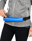 A person wearing a gray heathered zip-up jacket and black pants carries a bright blue waist bag with a black buckle while inserting keys into the front pocket.