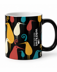A black ceramic mug featuring a colourful pattern of stylised New Zealand birds in yellow, teal, pink, and cream, with the text 'wild kiwi NEW ZEALAND' printed vertically on the side.