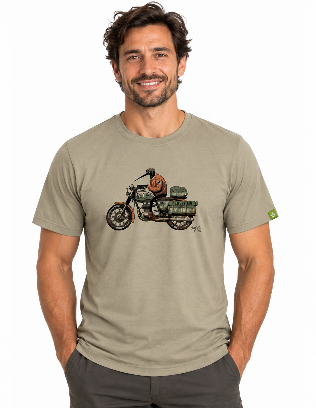 A man wearing a light khaki t-shirt with a vintage motorcycle graphic on the chest, paired with dark gray pants, against a plain white background.