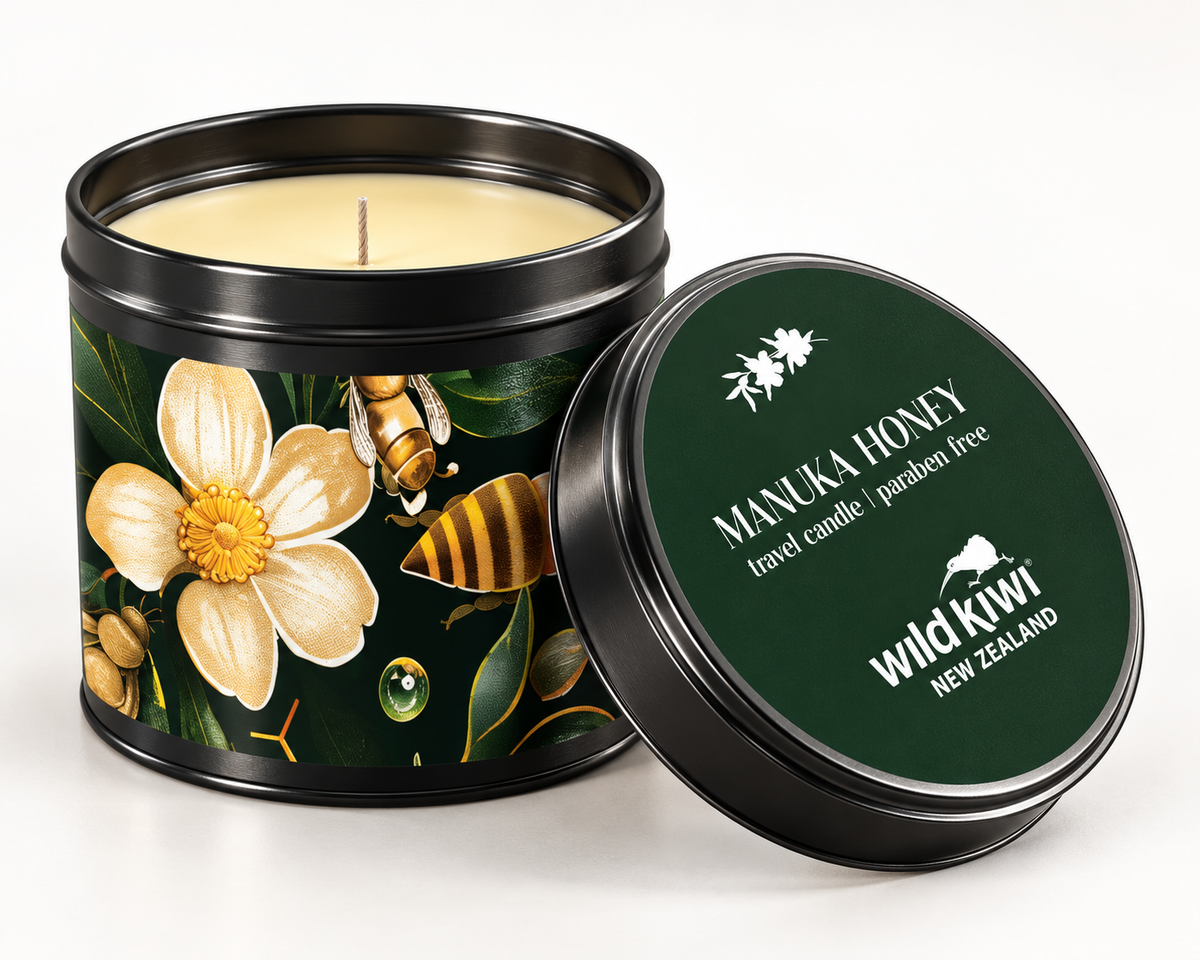 A black metal travel candle tin with a lid, shown open with cream-colored wax inside. The tin features a label with a white flower, green leaves, and a bee, while the lid is dark green with white text reading 'MANUKA HONEY travel candle' and 'wild kiwi NEW ZEALAND'.