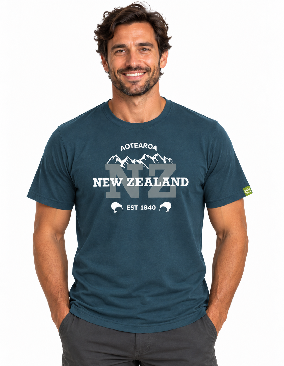 A smiling man wearing a dark teal blue t-shirt with a white graphic print that reads 'AOTEAROA NEW ZEALAND NZ EST 1840' with a mountain and rabbit design. The background is a clean white studio backdrop.