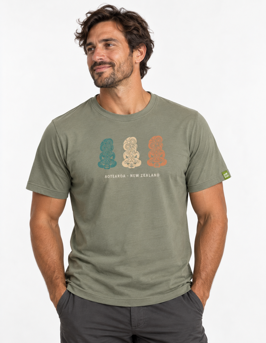 A man wearing an olive green t-shirt with a graphic of three Māori figures in teal, cream, and orange, with 'AOTAROA - NEW ZEALAND' printed below. The photo is taken against a clean white background.