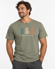 A man wearing an olive green t-shirt with a graphic of three Māori figures in teal, cream, and orange, with 'AOTAROA - NEW ZEALAND' printed below. The photo is taken against a clean white background.